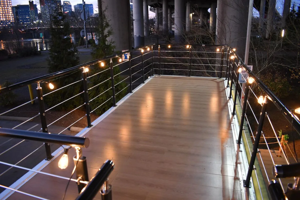 The rooftop deck with cable railings and string lights overlooking a city river